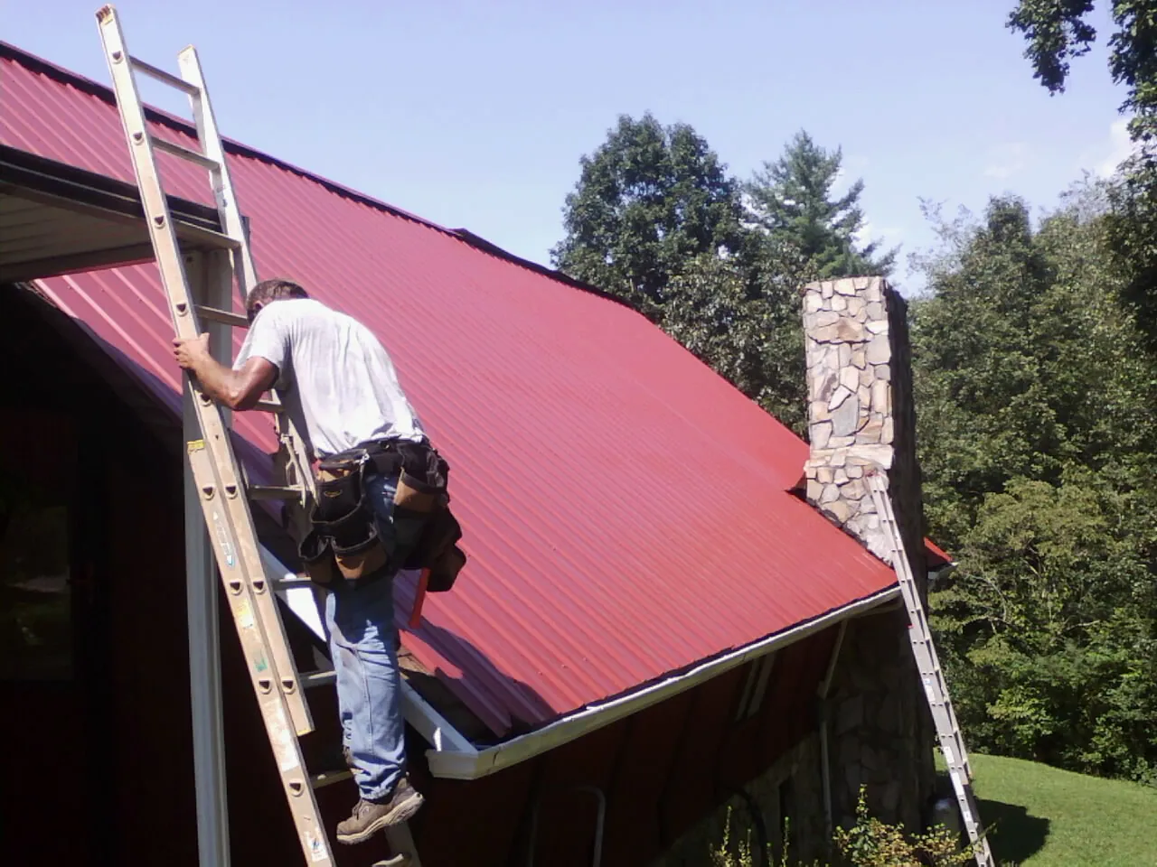 Roofer on a ladder beside a red metal roof and stone chimney — Soffit & Fascia Repair by Roofing Brothers in Boone Grove